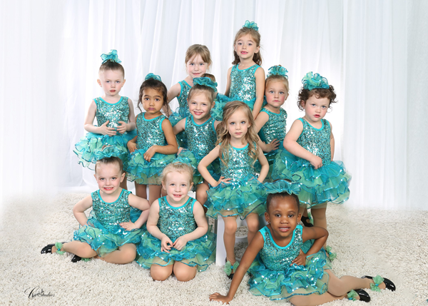 Group of young girls in teal sequined dance costumes, posing on a fluffy carpet in front of a white curtain.