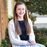 Woman with brown hair smiling in a car; light green top, beige car interior, blue window.