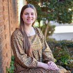 Woman with brown hair smiling in a car; light green top, beige car interior, blue window.
