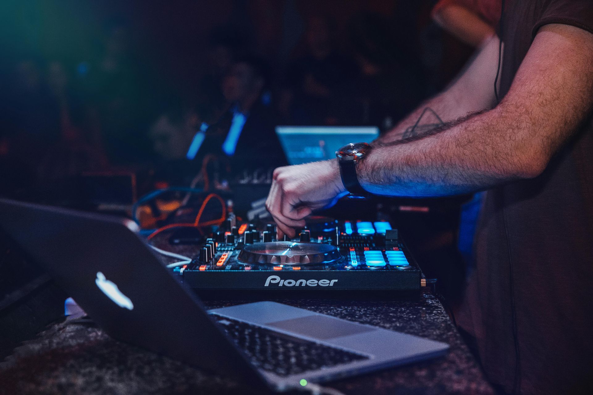 DJ op het werk, met een Pioneer-mixer, laptop, in een club. Armen met tatoeages, blauwe en donkere tinten.
