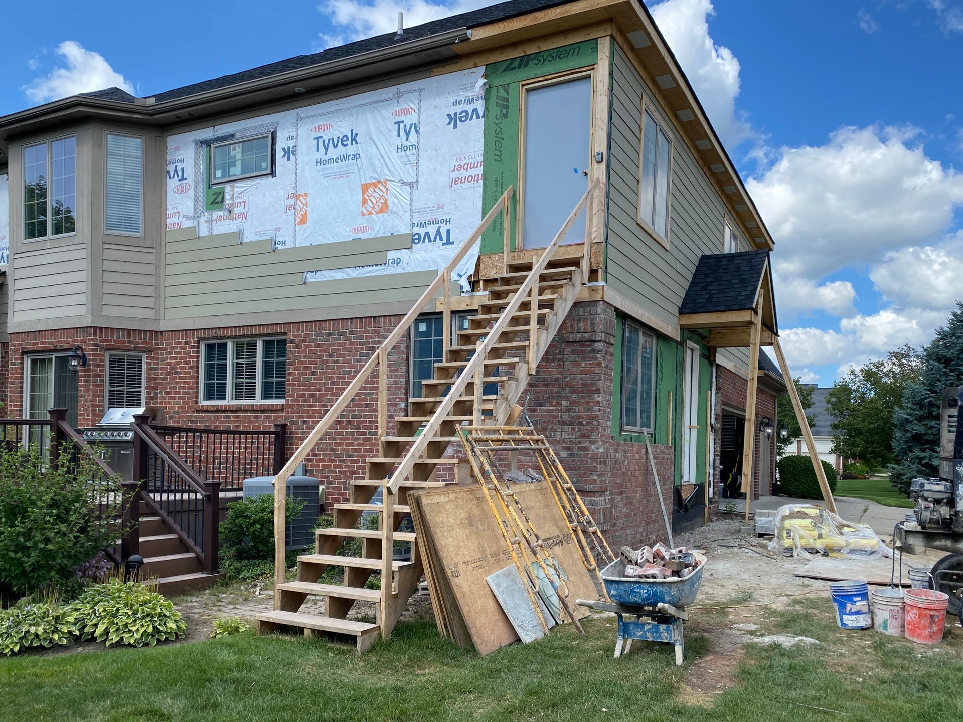 A two-story house under construction with exterior stairs. Brick, siding, and Tyvek wrap are visible.