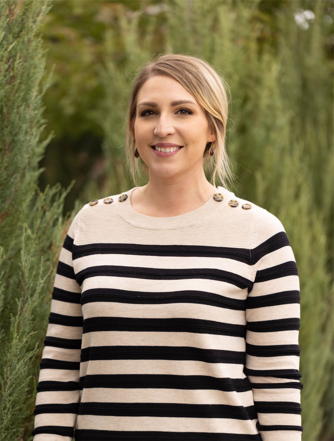 A woman in a striped sweater is standing in front of trees.