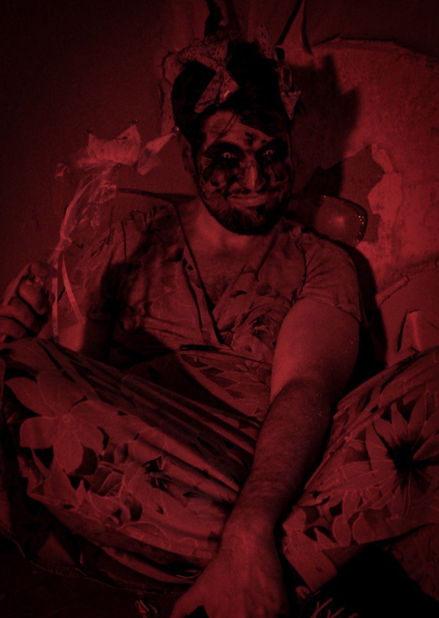 Man in a dimly lit, red-toned room, with dark markings on his face, seated in a tattered chair, looking forward.