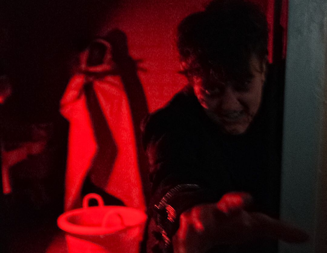 Person in dark clothing reaching out, lit by red light. Bucket and object on the wall visible.
