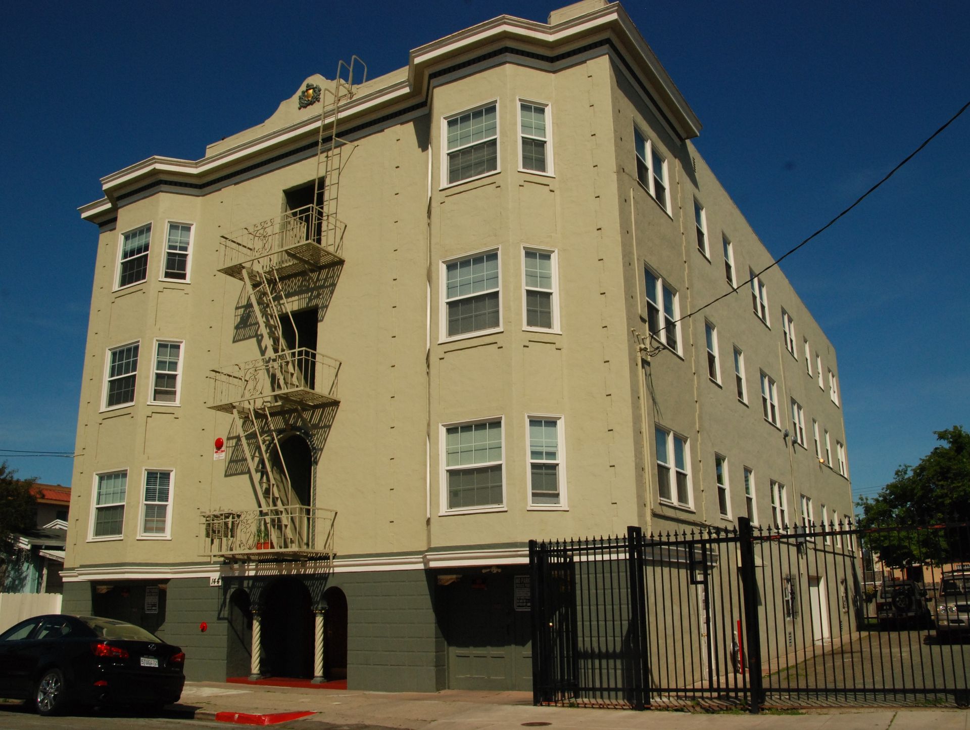 Beige four-story apartment building with fire escape and black fence on a sunny street corner