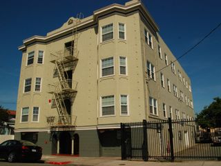 Beige four-story apartment building with fire escape and black fence on a sunny street corner