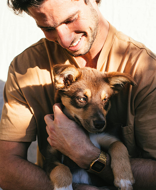 Man holding a puppy, both smiling. The man wears a watch and a tan shirt. Outdoor setting.