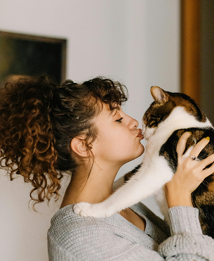 Woman holding a calico cat, about to kiss it. The woman has curly hair in a ponytail and is wearing a gray sweater.
