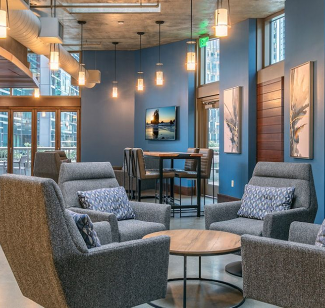 A modern lounge area with gray armchairs, a round table, and a dining table, set against blue walls with pendant lights.