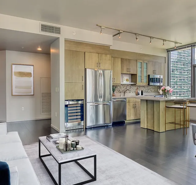 Modern open-plan kitchen and living room with stainless steel appliances, light wood cabinets, and gray flooring