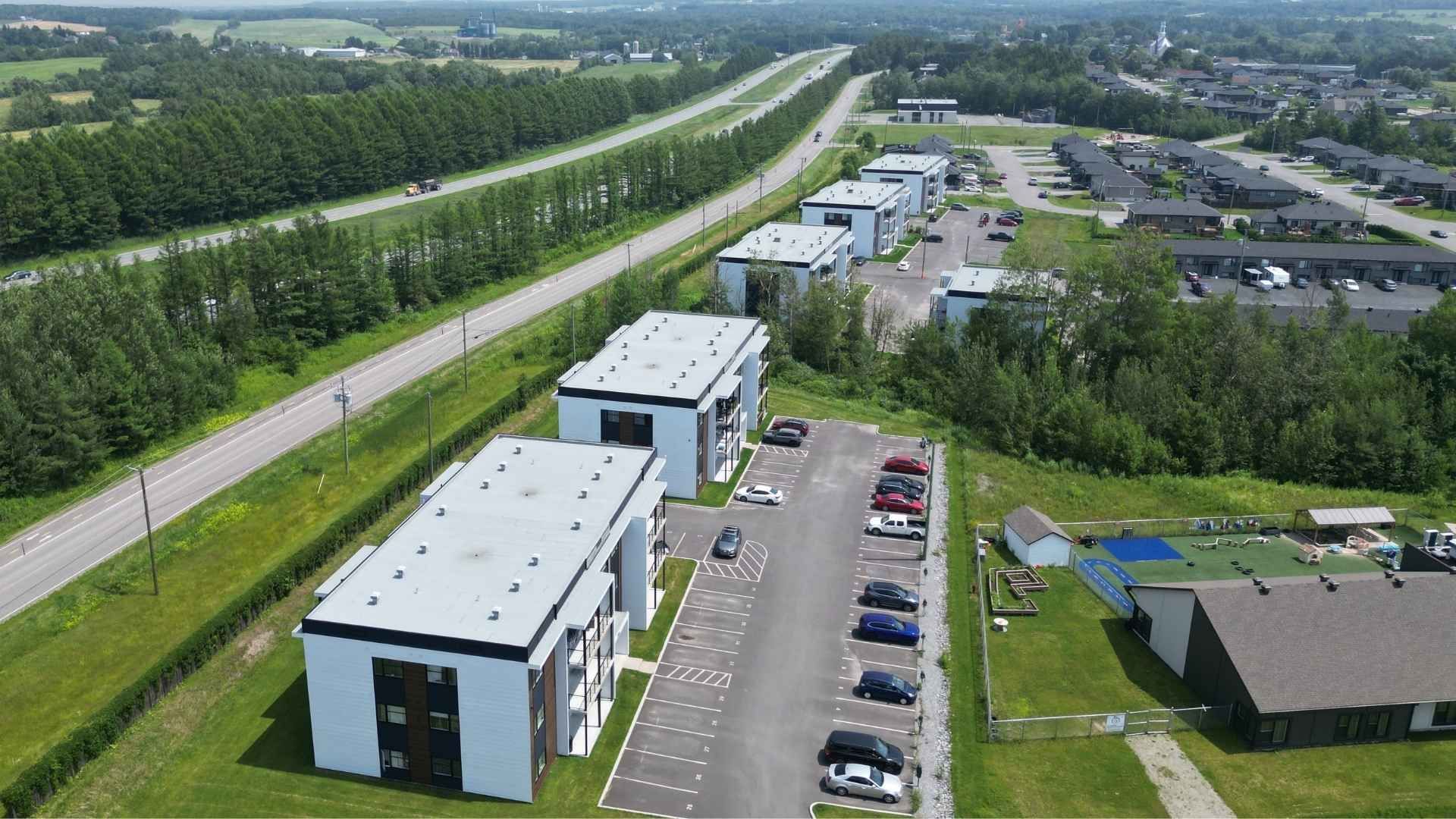 Vue aérienne de bâtiments modernes avec parking, à proximité d'une autoroute et d'arbres verdoyants.