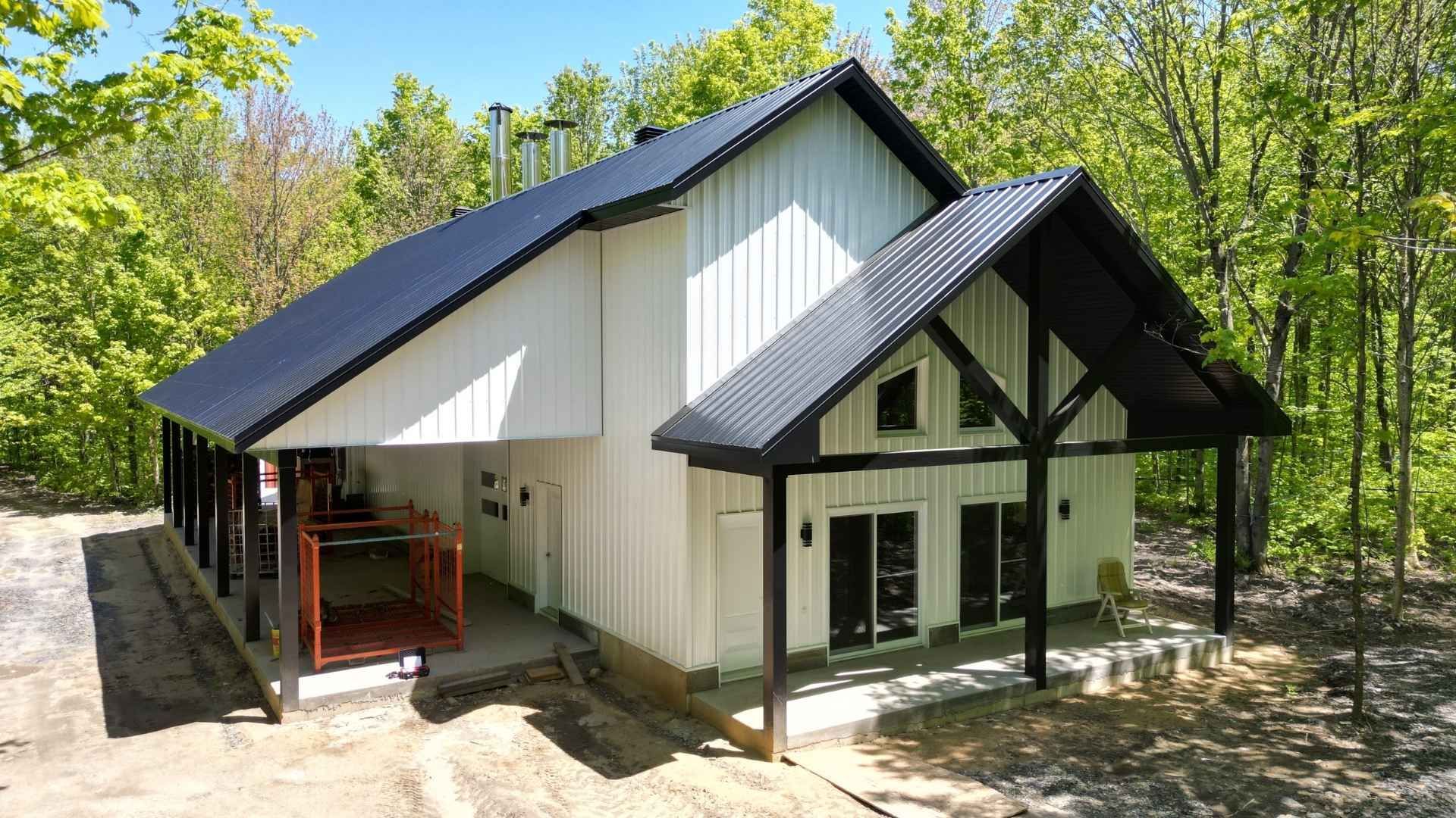 Cabane à sucre moderne blanche avec toit et boiseries noires, entourée d'arbres.