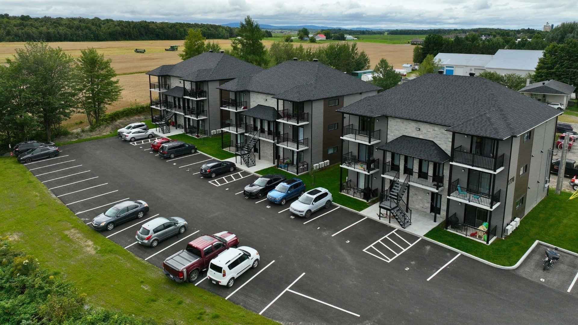 Immeubles d'appartements de trois étages avec balcons et parking, situés à proximité d'un champ.