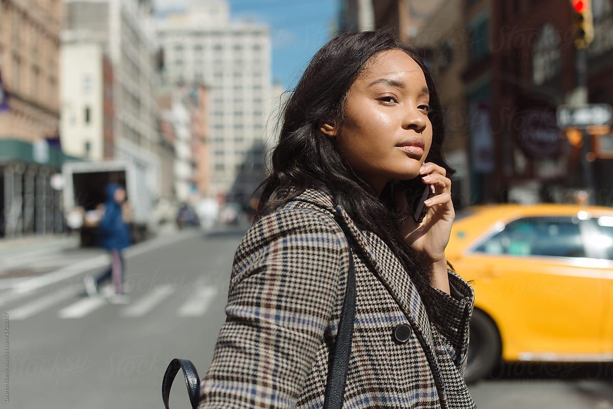 A woman on her phone crossing the street in a city with a yellow taxicab visible behind her.