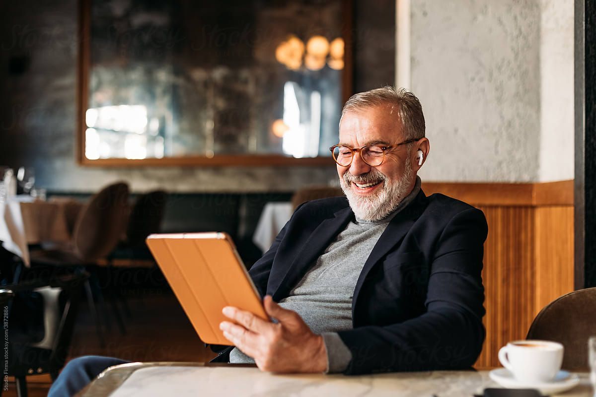 A bearded man smiling while seated in a coffee shop, reading something on his iPad.