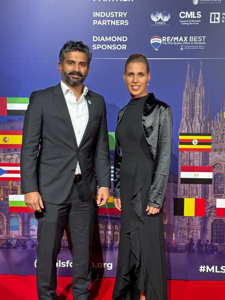 Mathew Kallumadil and Marion Wieler pose at event with international flags. Mathew in suit, Marion in black dress and jacket.