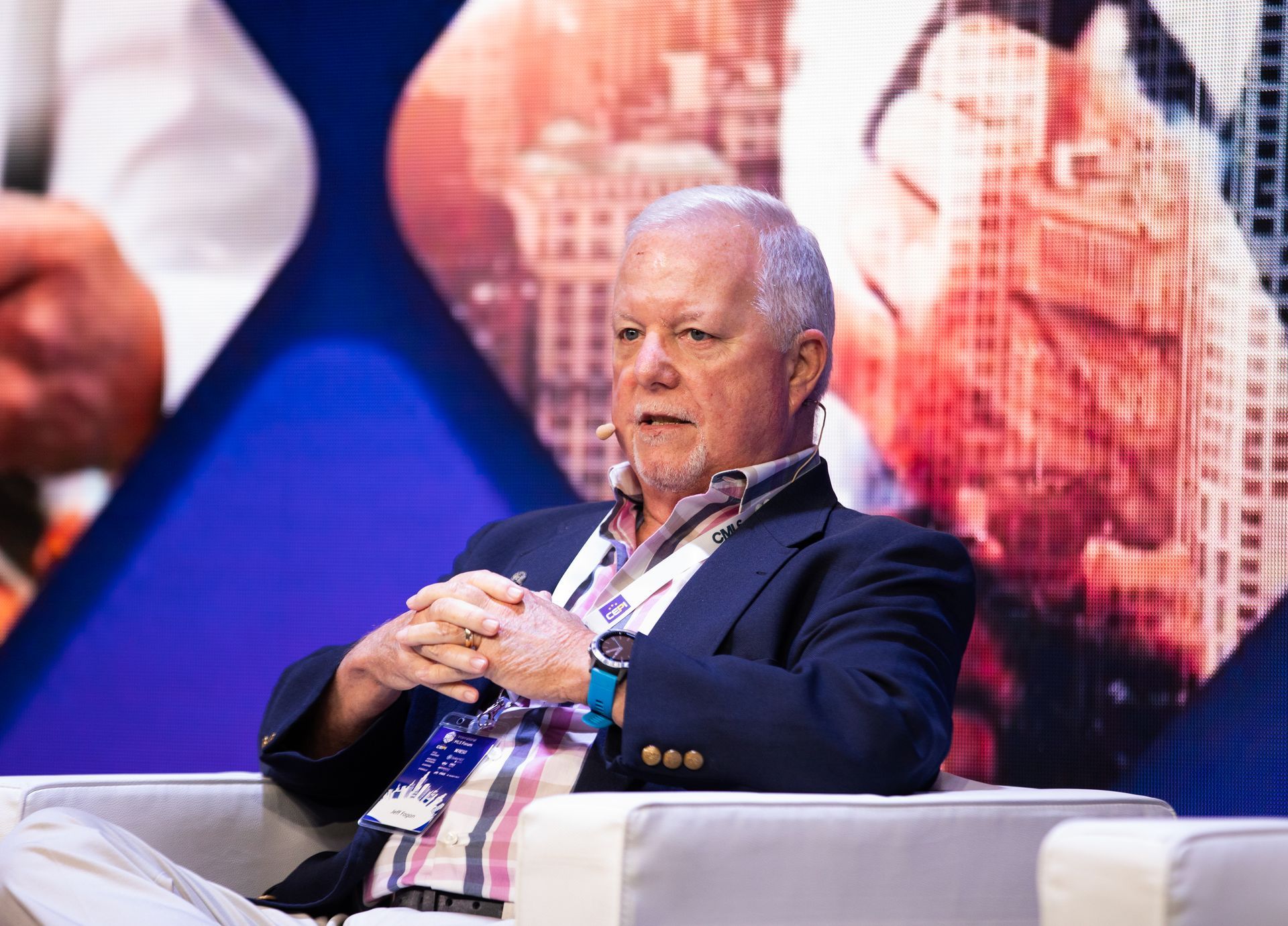 Jeff Fagan seated, speaking at an event, wearing a blazer over patterned shirt; city backdrop.