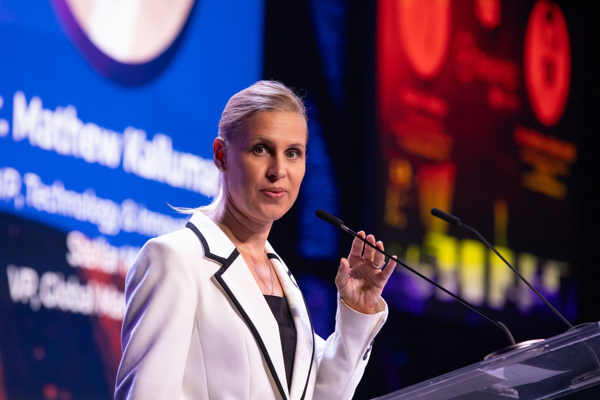 Marion Weiler in a white jacket speaking at a podium with a microphone. Large screen in the background.
