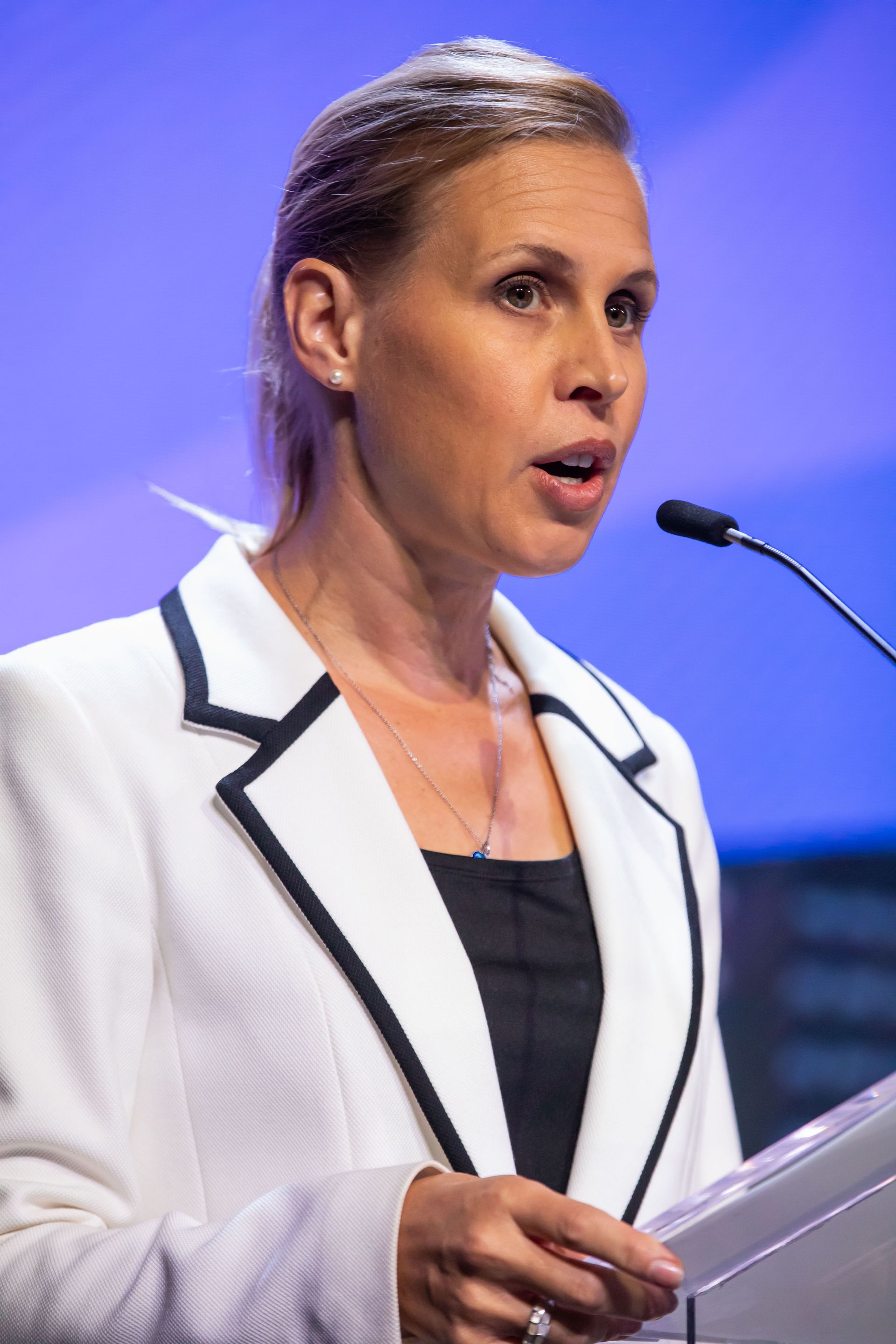 Marion Weiler speaking at a podium, wearing a white blazer with black trim, holding notes.