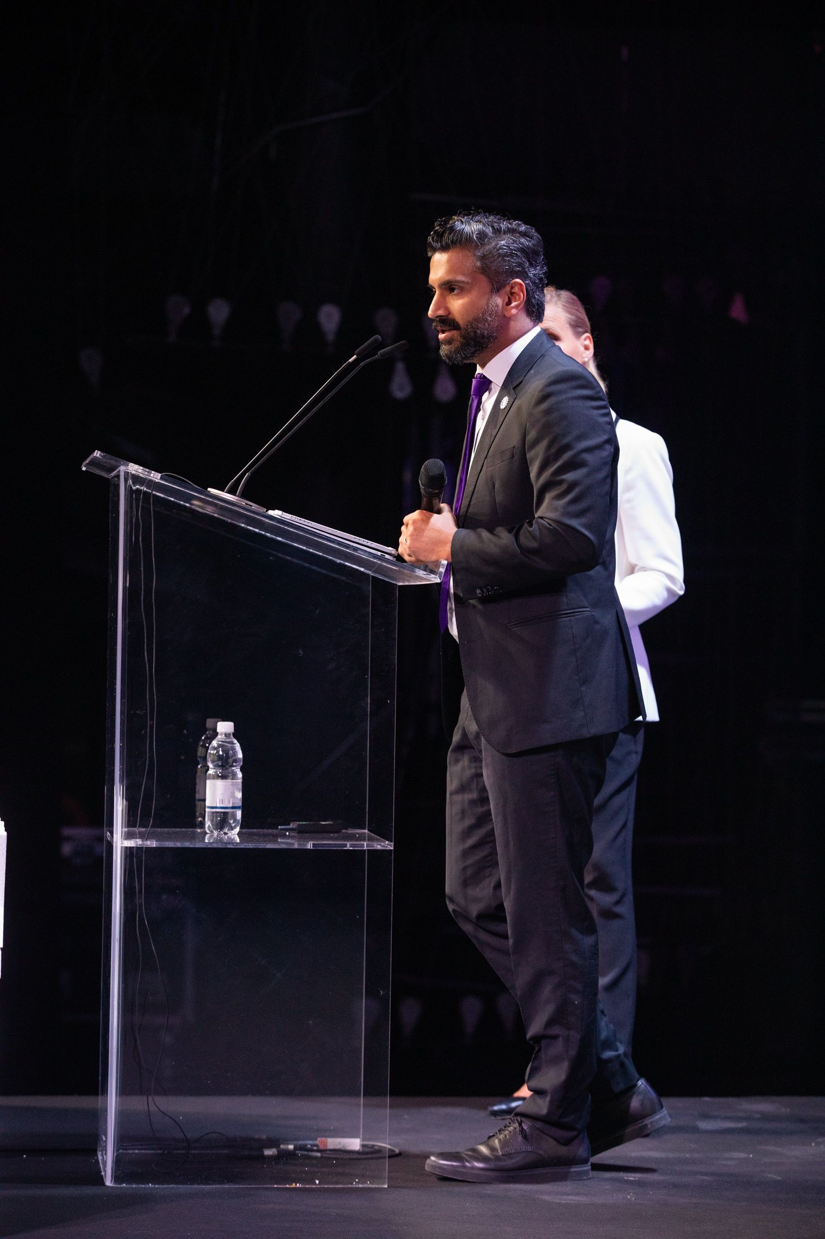 Mathew Kallumadil in a suit speaking at a clear podium with a microphone, Marion Weiler stands nearby. Dark stage.