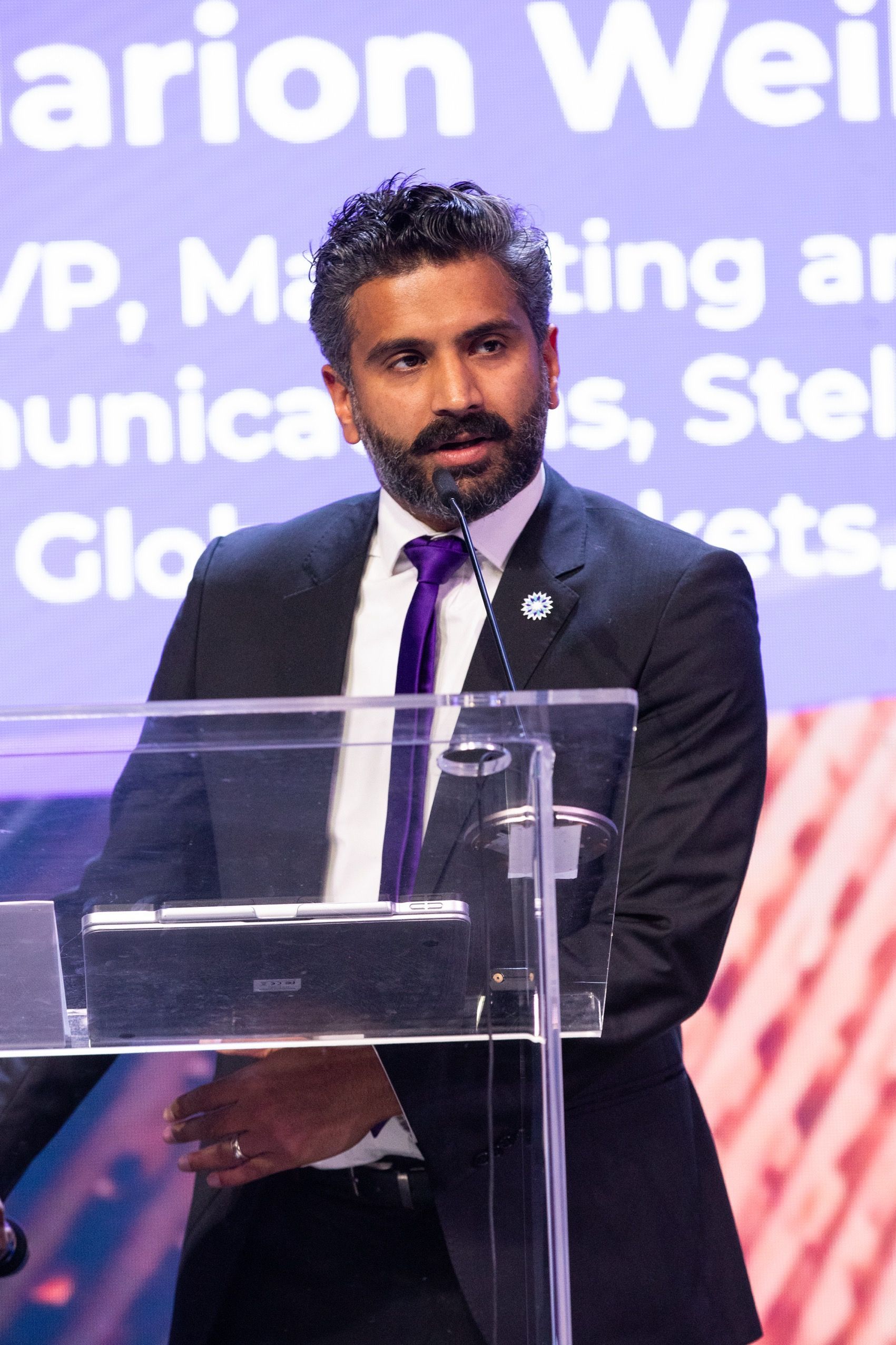 Mathew Kallumadil in a suit speaks at a podium; purple tie, dark hair, behind a clear barrier.