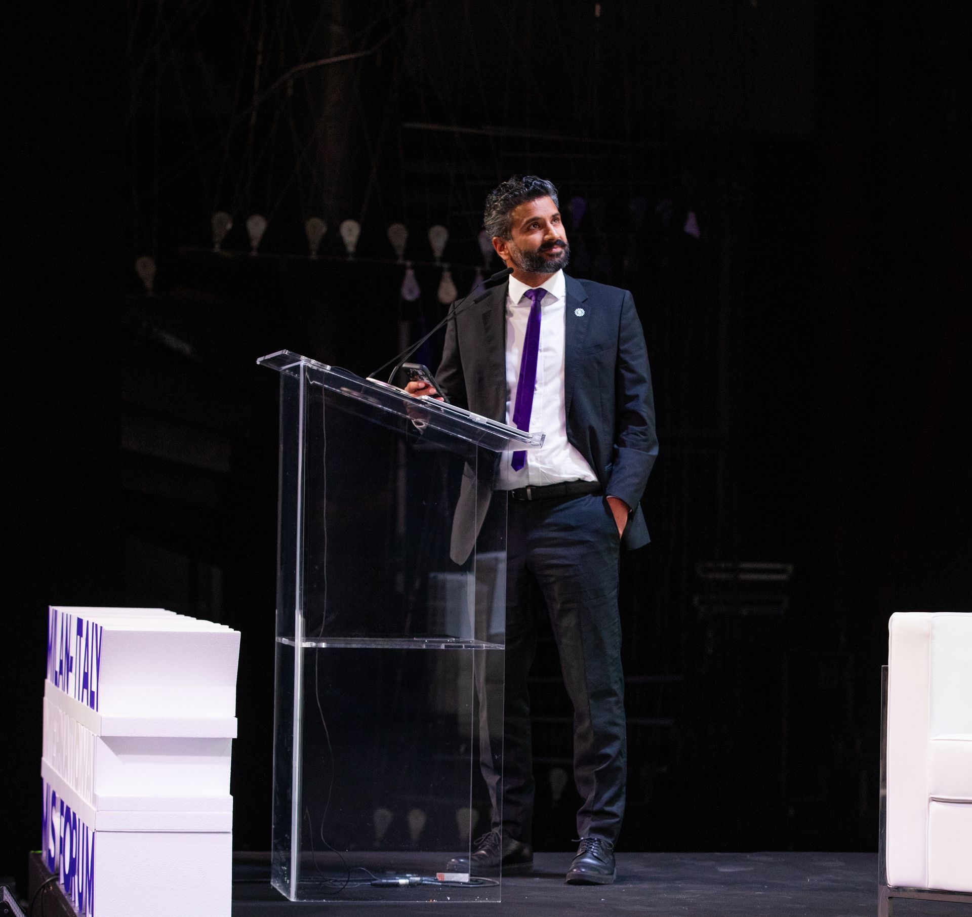 Mathew Kallumadil in a suit at a podium, looking to the side. Stage setting with boxes and lights.