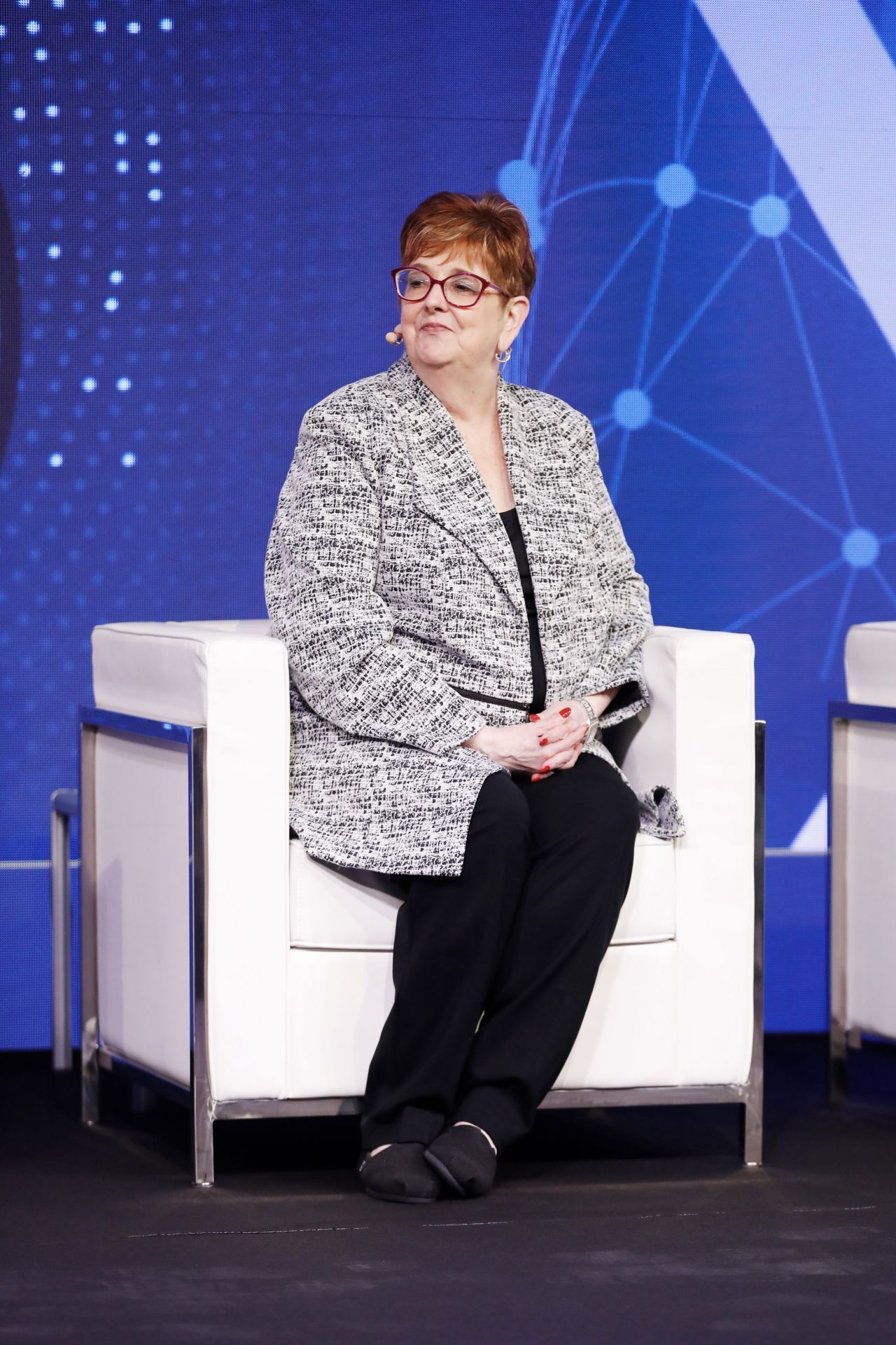 Merri Jp Cowen in black-and-white jacket sits on a white chair, hands clasped, on stage.