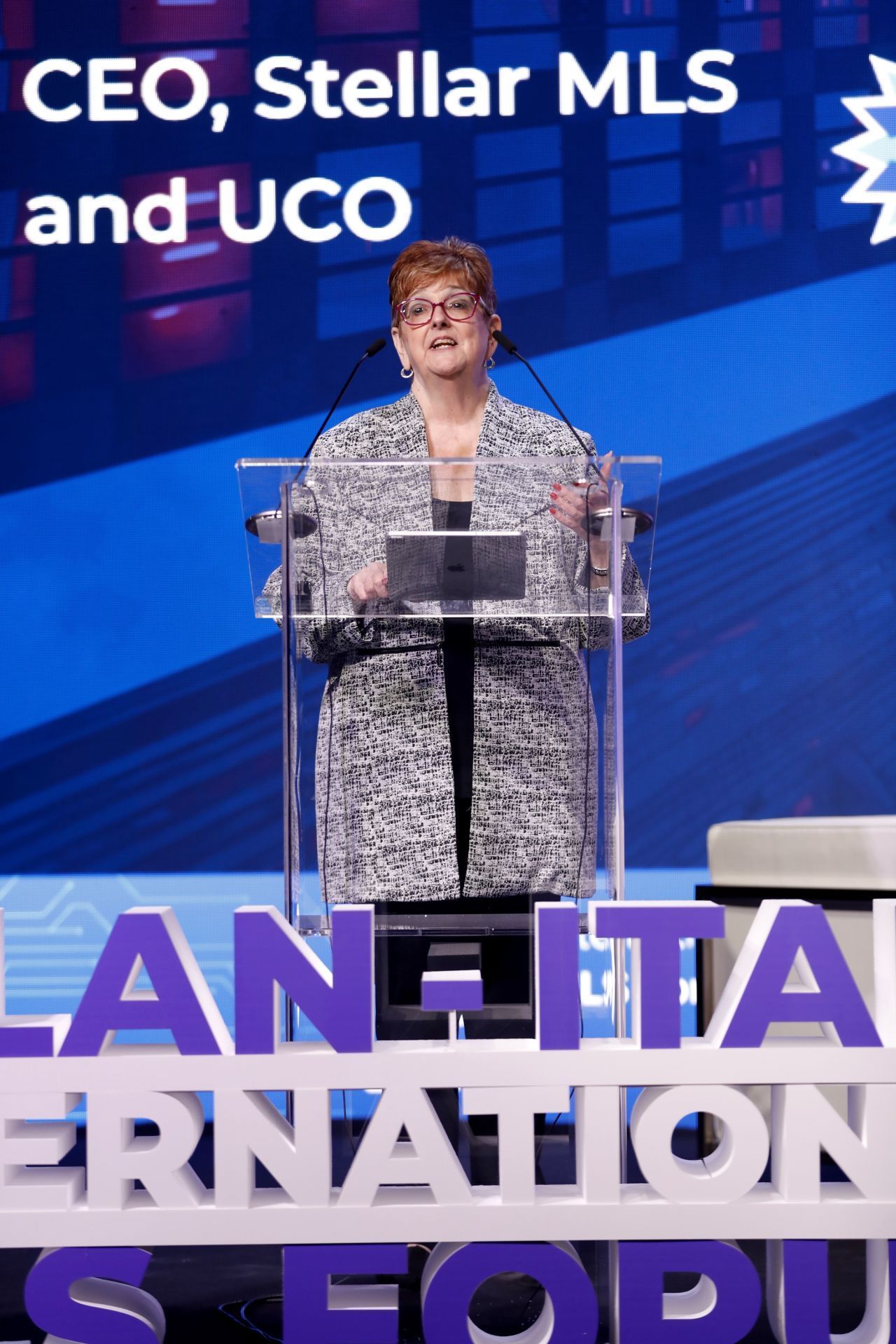 Merri Jo Cowen speaking at a conference, behind a podium. Text on stage: PLAN-ITALIA INTERNATIONAL FORUM.