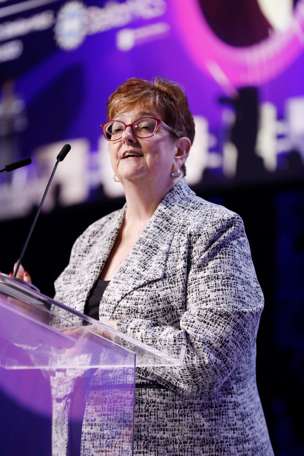 Merri Jo Cowen speaking at a podium, wearing a patterned jacket, lit on a stage.