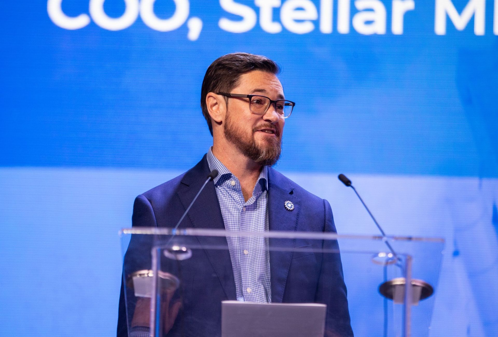 Shayne Fairley speaking at a podium, wearing glasses, blue blazer and patterned shirt, with a laptop in front of him.