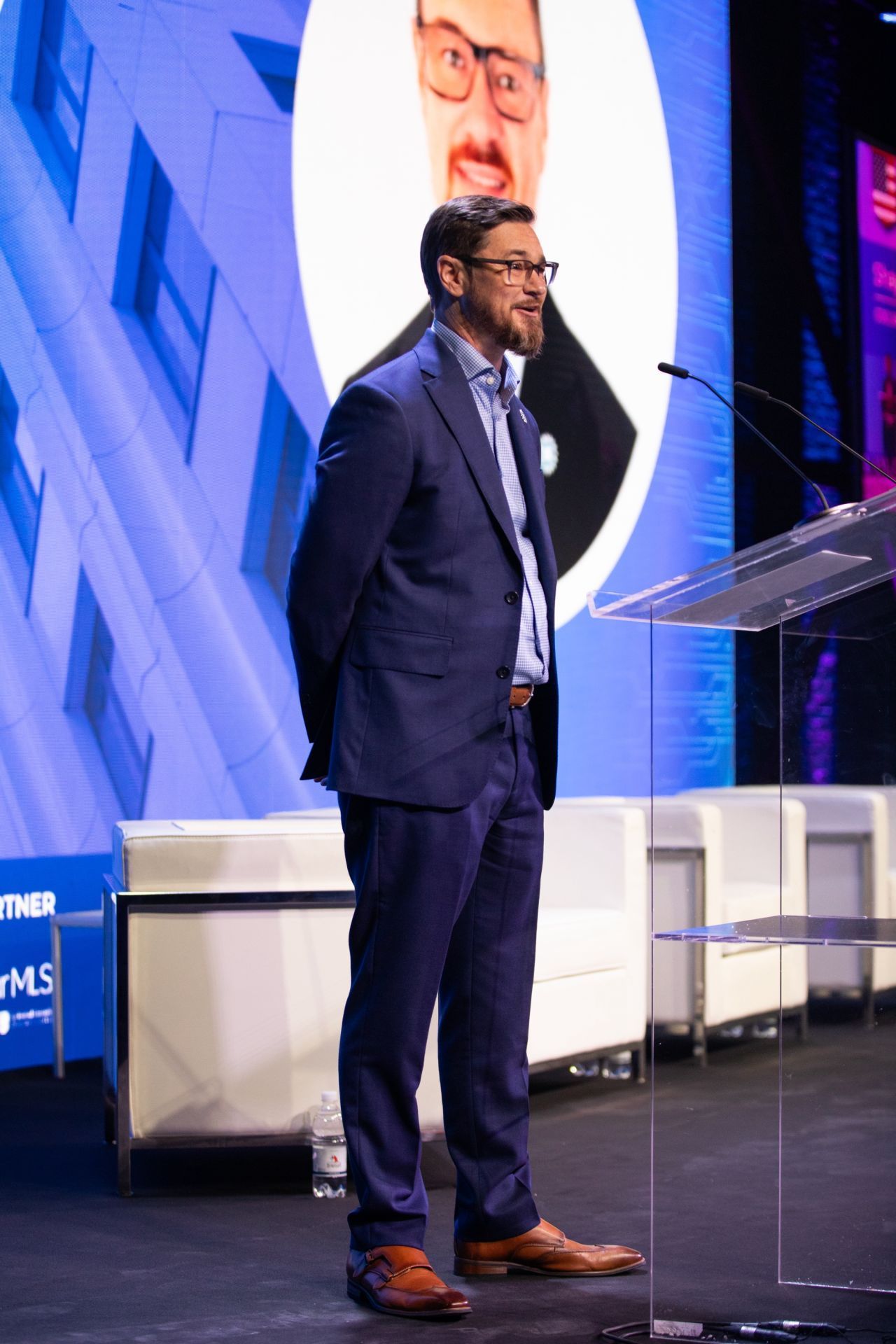 Shayne Fairley in blue suit stands at a podium, speaking into a microphone with a large image behind him.