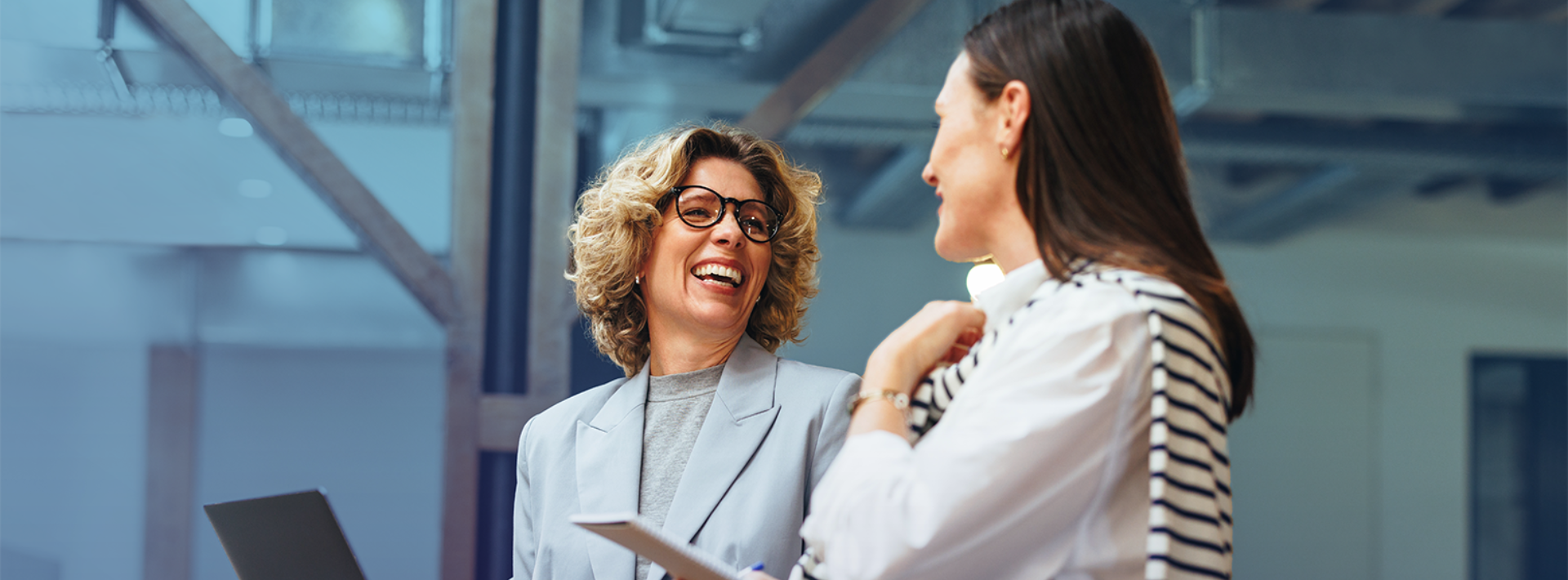 Two professionals in an office setting smile and converse while holding a laptop and tablet.