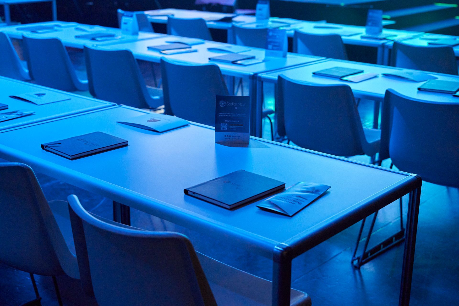 Empty tables with chairs arranged in rows, illuminated by blue light in a modern venue.