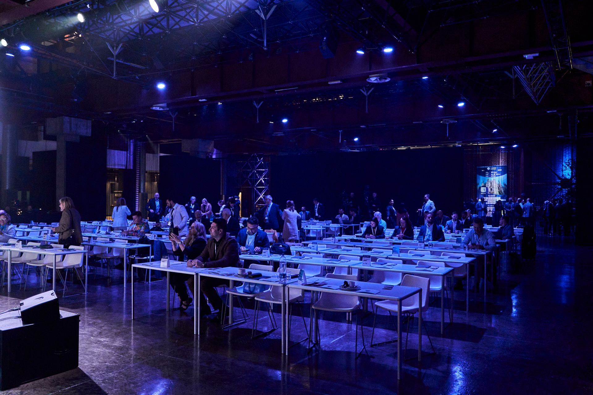 Large room with tables, attendees, and blue lighting; looks like a conference or event.