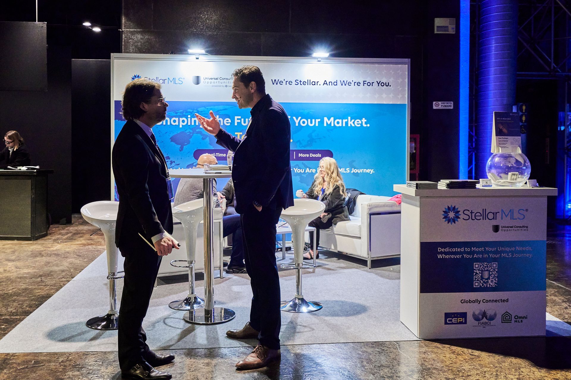 Two men converse at a conference booth. White and blue backdrop with text, people seated, silver tables.
