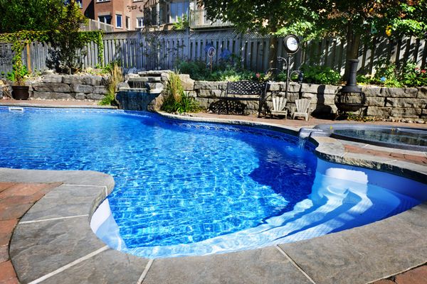 A blue kidney-shaped swimming pool with built-in steps, surrounded by stone coping and a landscaped backyard.