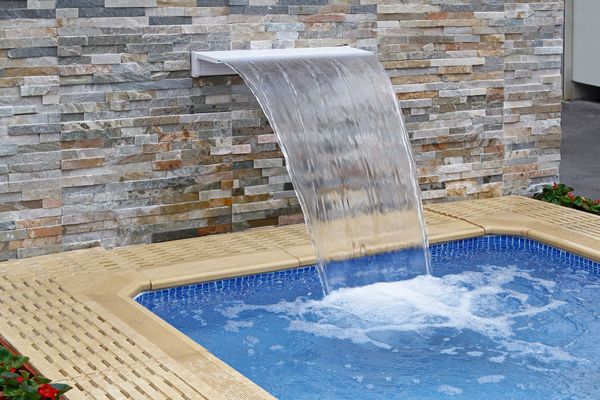 A water feature cascading from a stone wall into a blue-tiled swimming pool.