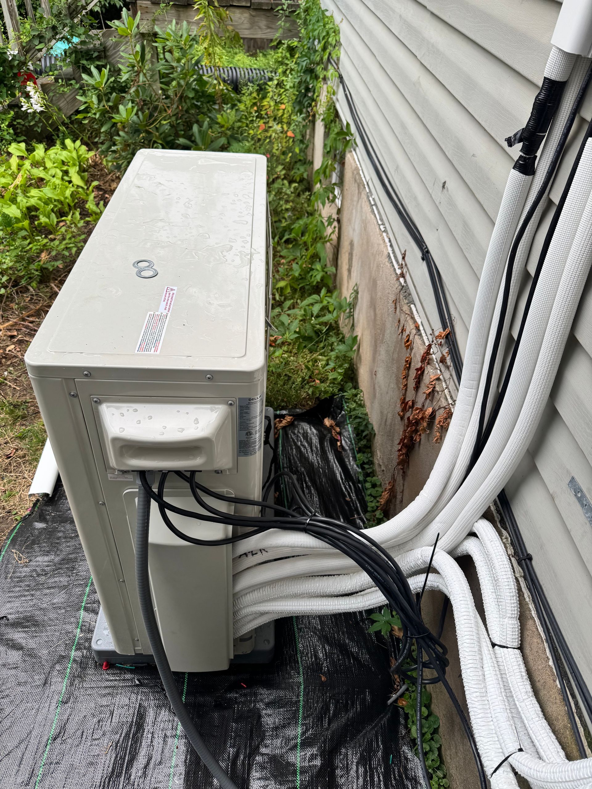 Outdoor air conditioning unit beside a building, with multiple insulated white tubes running along the wall.