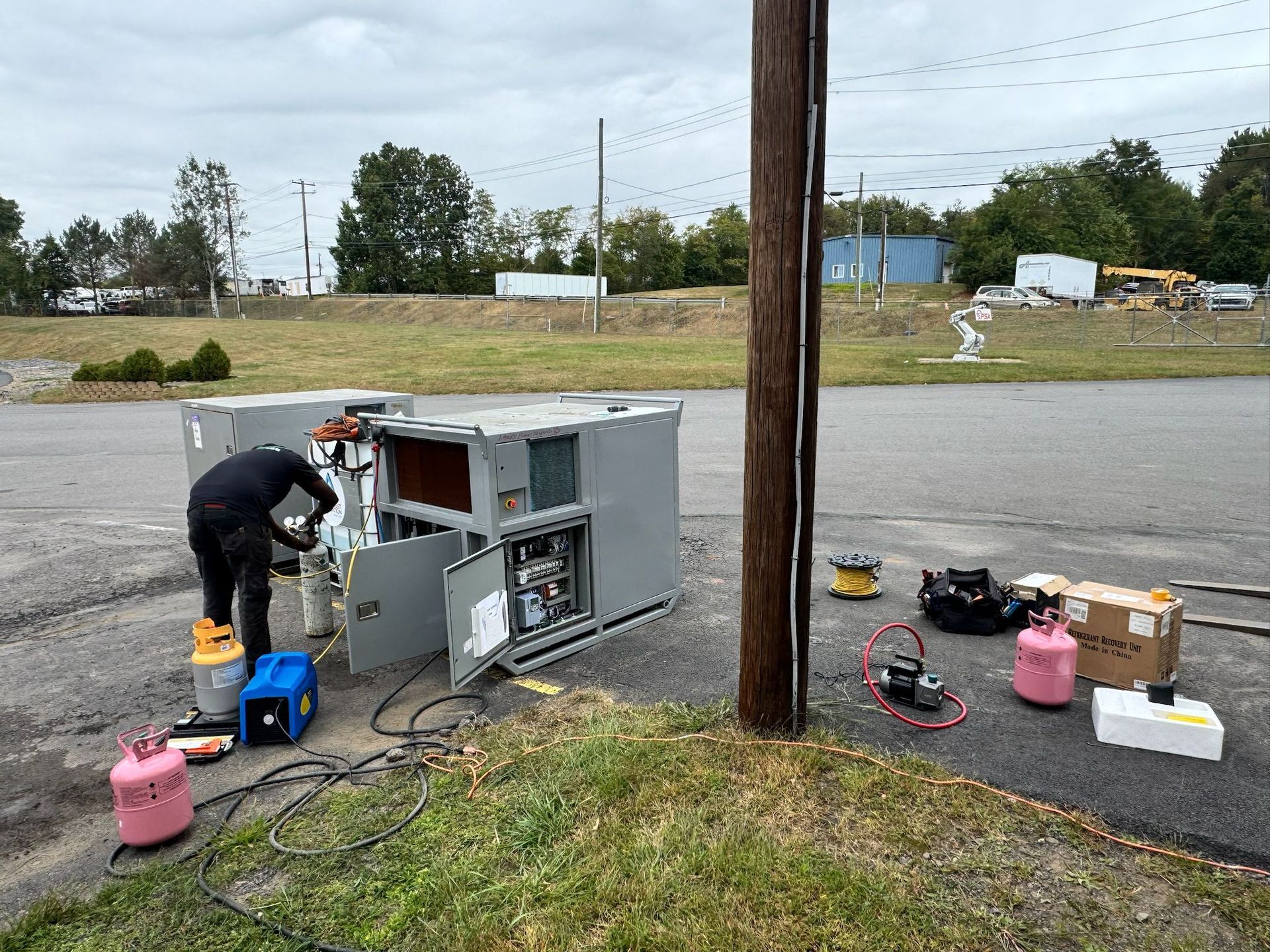 HVAC technicians working on two large gray units outdoors, next to a pole. Equipment and tools are nearby.