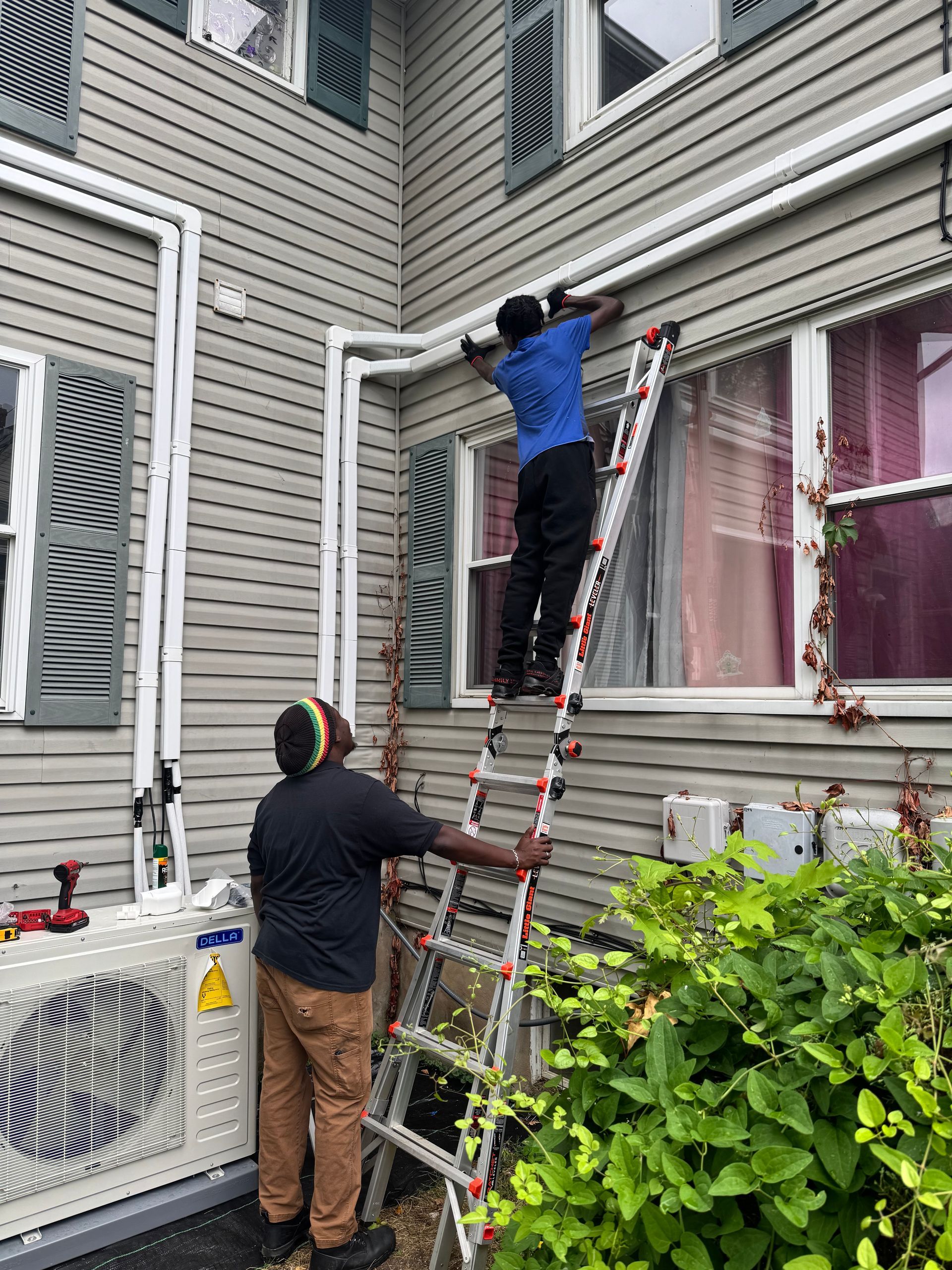 Two workers installing white pipes on a gray house exterior. One on a ladder, the other at ground level.