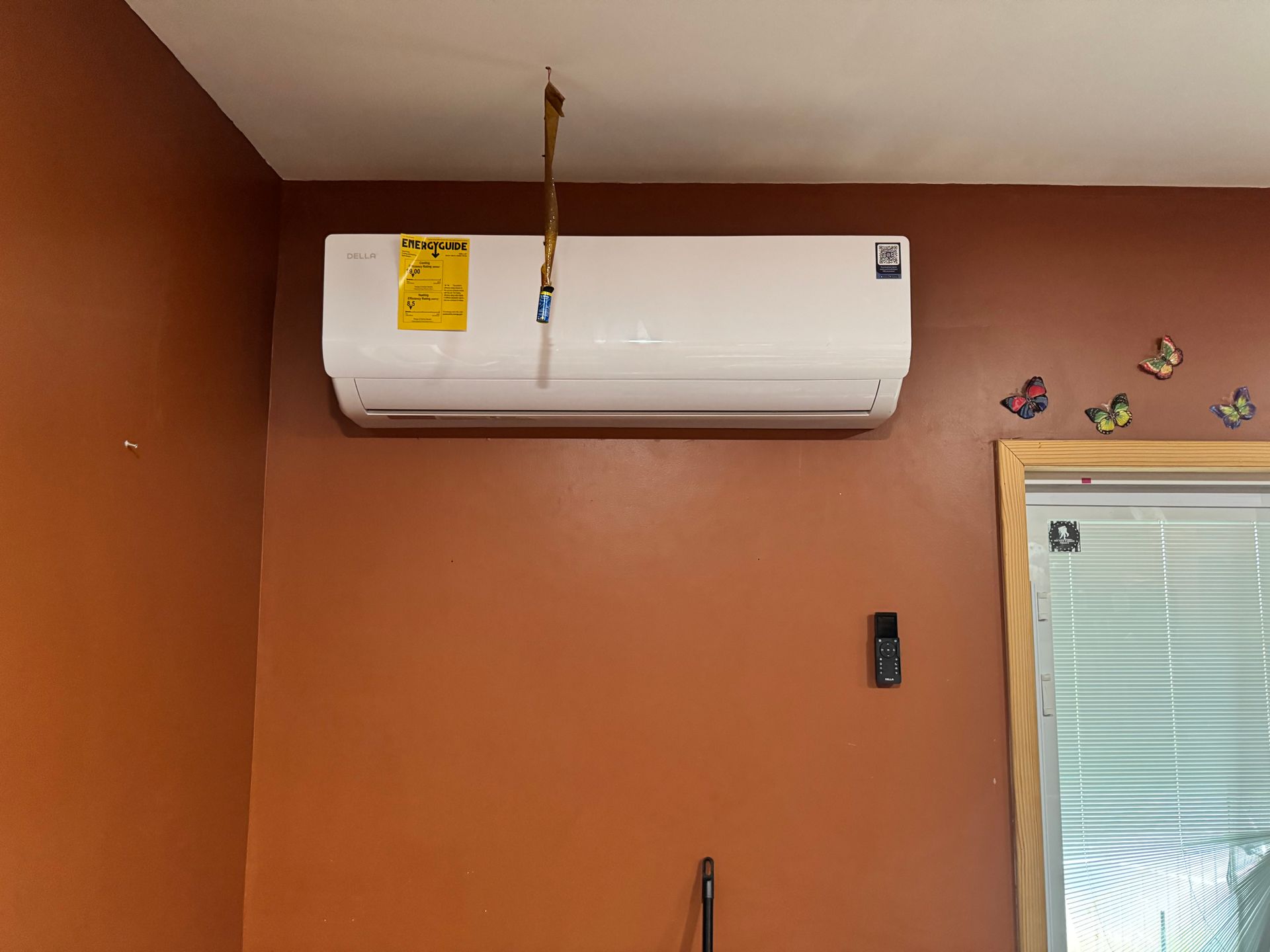 White wall-mounted air conditioner on a brown wall with a remote control, near a window with blinds.