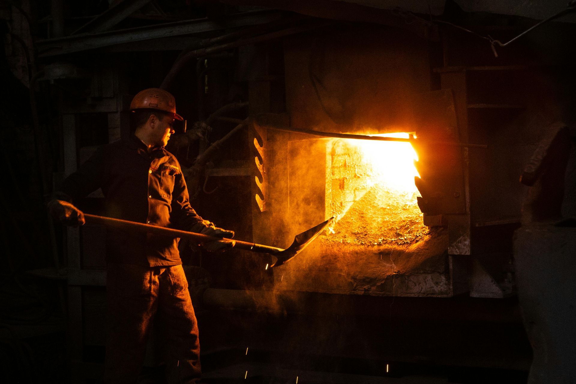 Worker using a long tool to tend to a fiery furnace in a dimly lit industrial setting.