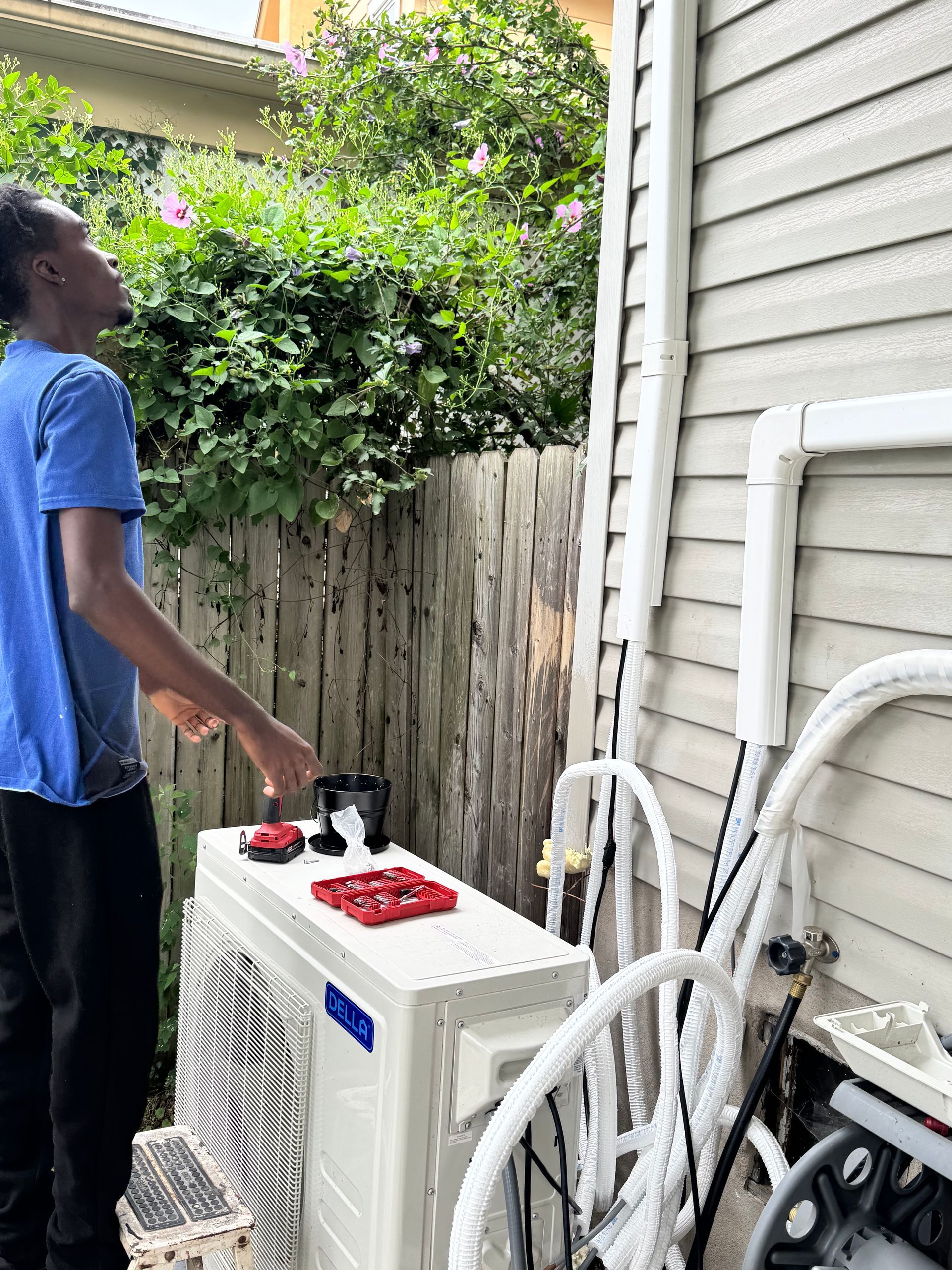 Person working on an air conditioning unit outside near a wooden fence and building with white siding.
