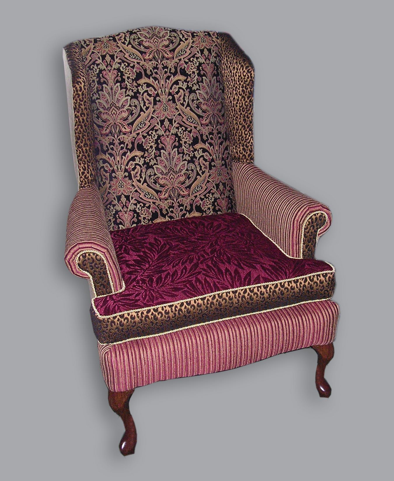 An ornate wingback chair with dark patterned fabric, a deep burgundy seat, and fringe trim, set on cabriole wood legs.