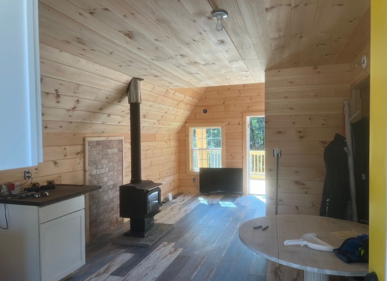 Interior view of a cabin with wood walls and ceiling, wood-burning stove, and a dark floor.