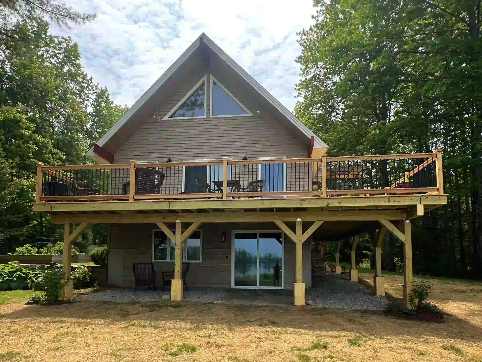 Two-story house with a large deck, surrounded by trees. Tan siding, angled roof, and light wood deck.