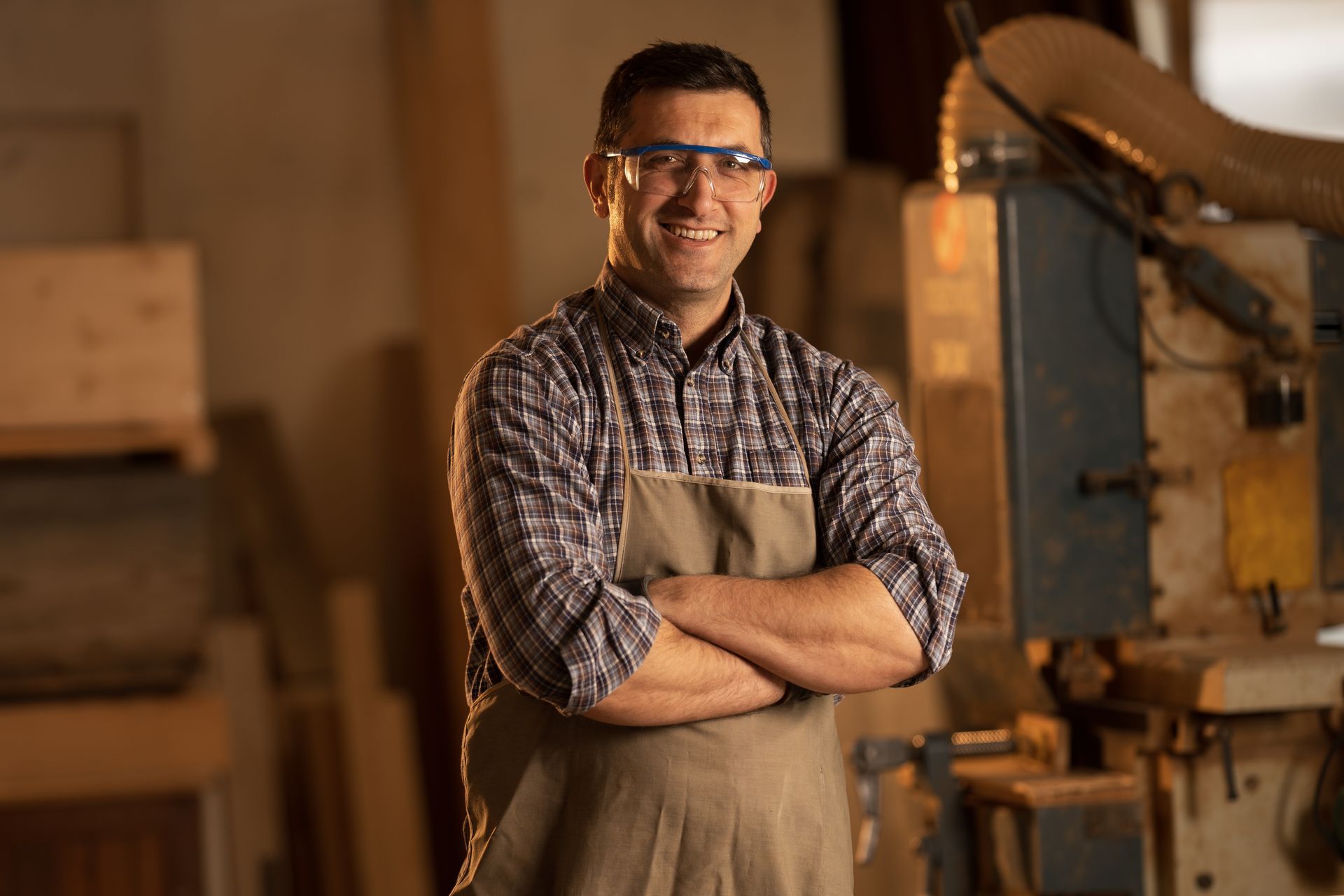 Man with arms crossed, wearing safety glasses and apron, in a wood shop, smiling.