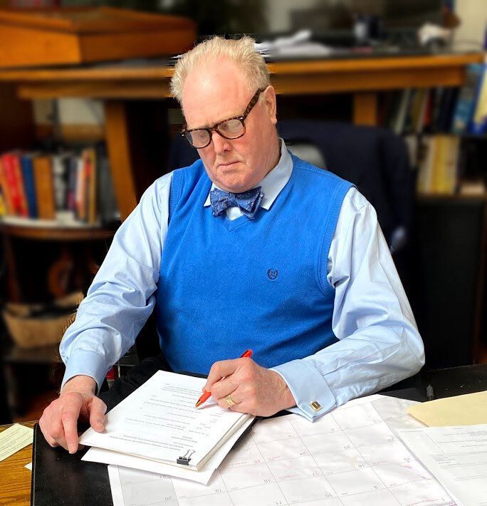 A man wearing glasses and a blue vest is sitting at a desk