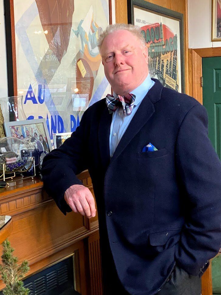 A man in a suit and bow tie is standing in front of a fireplace.