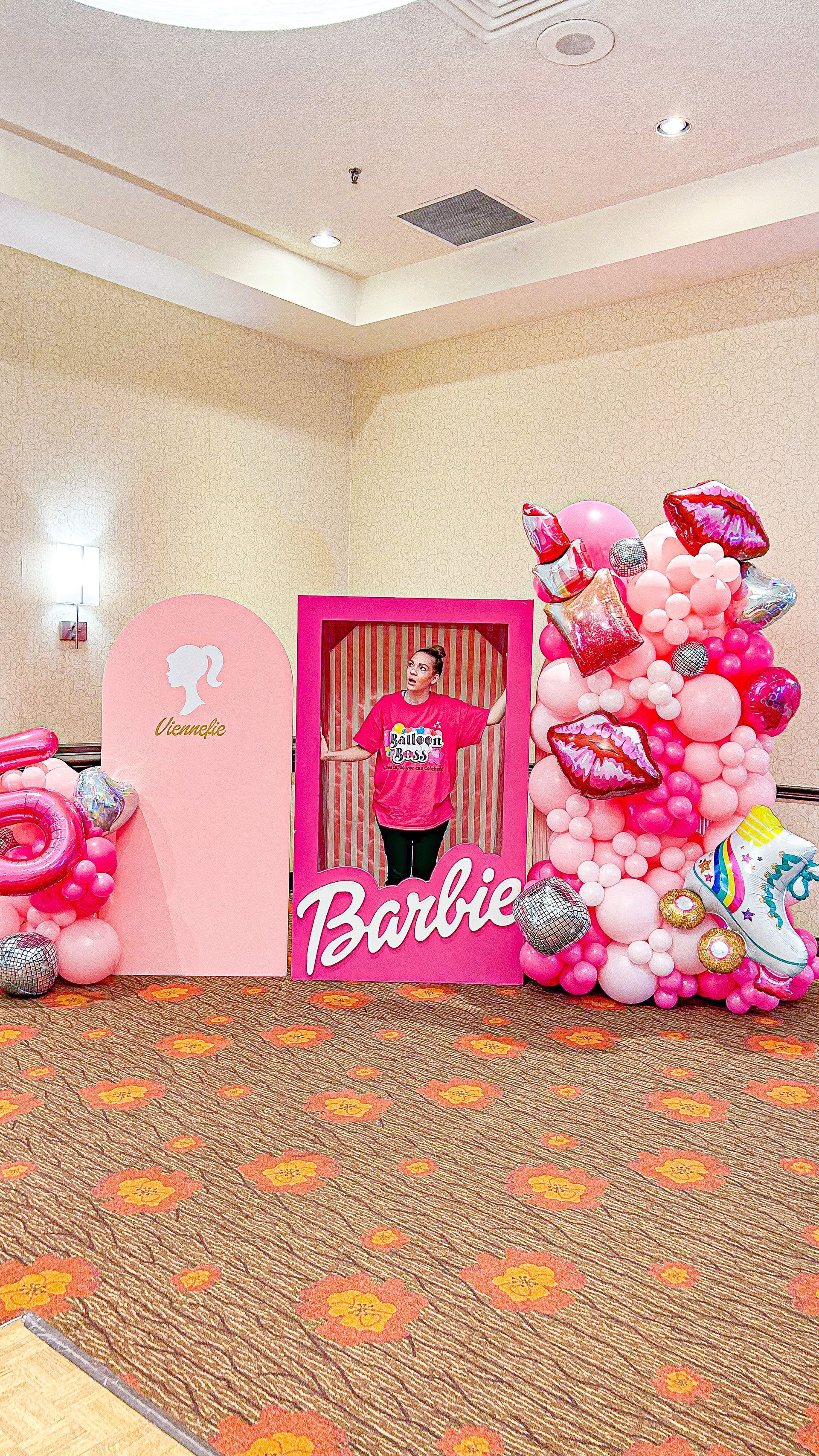 A woman is standing in front of a barbie sign surrounded by pink balloons.